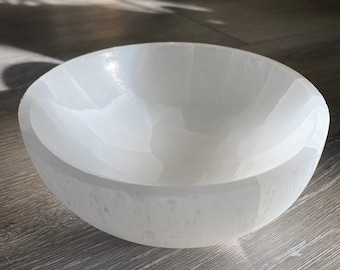 May include: A white, round bowl with a smooth, polished surface. The bowl is made of a translucent material, and the light reflects off its curved shape. The bowl is sitting on a wooden surface.
