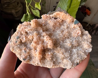 May include: A close-up of a natural mineral specimen. The stone features a mix of peach, cream, and white crystalline structures. Small patches of dark green and black are visible on the surface. The overall texture is rough and uneven.