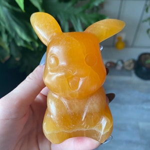 May include: A carved orange-yellow Pikachu figurine. The figurine has large ears, round cheeks, and a smiling expression. The sculpture is held in a hand, with a blurred background of green foliage.