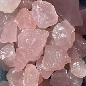 May include: A collection of raw rose quartz stones. The stones vary in size and shape, with a pale pink hue and a rough, textured surface. The image showcases the natural beauty of the crystals, perfect for collectors or those interested in healing stones.