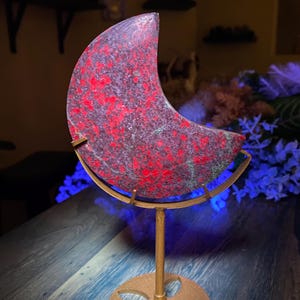 Puede incluir: Objeto decorativo en forma de media luna con un patrón moteado rojo y gris, montado sobre un soporte de metal dorado. El soporte tiene una base circular y un tallo delgado. El objeto se exhibe sobre una superficie de madera oscura.