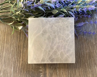 May include: A square, pale grey selenite crystal charging plate with a cracked, textured surface. The plate is surrounded by purple and green faux lavender sprigs, set against a wood-grain background.