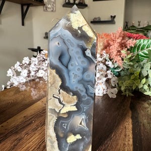 May include: A tall, polished agate crystal point with a flat base and a pointed top. The stone features swirling patterns of gray, blue, beige, and yellow. The crystal is displayed on a wooden surface.