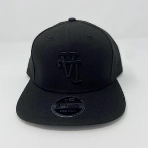 May include: A solid black baseball cap featuring a tonal embroidered logo on the front with the letters V and I intertwined. The cap has a flat brim and a circular sticker on the top of the brim that reads New Era 9FIFTY SNAPBACK.