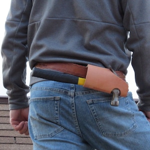 May include: A person wearing a gray sweatshirt and blue jeans with a brown leather hammer holder attached to their belt.