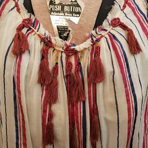 Puede incluir: Top a rayas color crema con rayas verticales rojas y azules. La parte superior presenta un escote en V adornado con adornos de cuentas y borlas rojas colgantes. Una etiqueta dice "PUSH BUTTON ADJUSTABLE DRESS FORM".