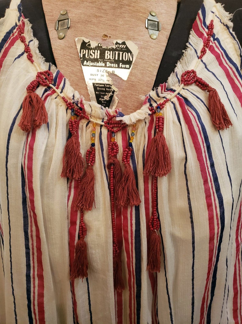 Puede incluir: Top a rayas color crema con rayas verticales rojas y azules. La parte superior presenta un escote en V adornado con adornos de cuentas y borlas rojas colgantes. Una etiqueta dice "PUSH BUTTON ADJUSTABLE DRESS FORM".