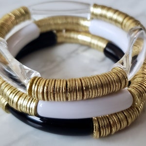 May include: Three stacked bracelets, one clear, one black and white, and one gold with small round discs.