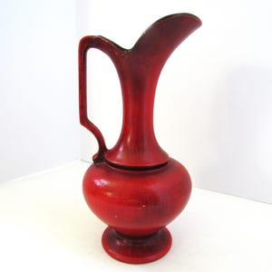 May include: A vibrant red pitcher featuring a rounded base, a slender neck, and a curved handle. The surface displays a textured, mottled finish with darker red shading throughout. The pitcher has a wide, flared spout at the top and stands on a circular pedestal base.