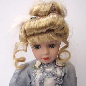 May include: A porcelain doll with blonde hair styled in an updo. She is wearing a light blue dress with white lace trim and floral accents.