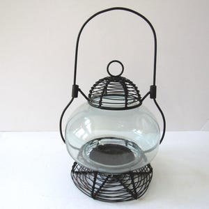 May include: A decorative glass lantern with a round, clear glass body and a black wire cage design. The lantern has a black wire handle and base, suitable for candles or small lights. The overall design is vintage and rustic.