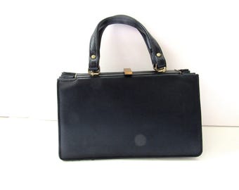 Könnte beinhalten: Eine schwarze Vintage-Handtasche mit einer strukturierten rechteckigen Form. Sie verfügt über einen geschwungenen Griff, goldfarbene Beschläge und einen Schnallenverschluss. Die Tasche besteht aus einem glatten Material, möglicherweise Leder, und ist vor weißem Hintergrund platziert.