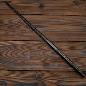 May include: A black wand with intricate carved designs. The wand is long and slender, with detailed patterns along its length. The wand is set against a dark wood background.