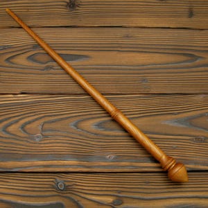May include: A handcrafted wooden wand with a light brown finish. The wand features a tapered design with a detailed handle and a rounded tip. The wand is set against a wooden background.