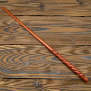 Puede incluir: Una varita de madera hecha a mano con un rico acabado marrón. La varita presenta intrincados detalles tallados a lo largo de su longitud, incluido un patrón de diamantes y un diseño en espiral. La varita mide aproximadamente 36 cm de largo.
