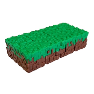 May include: A rectangular block toy with a pixelated design, reminiscent of a video game. The top is green, simulating grass, while the sides are brown, representing earth. The toy is designed to resemble a block from the popular game.