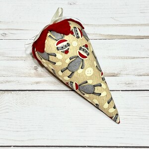 May include: A cone-shaped fabric ornament with a red felt top and a beige fabric base. The fabric is printed with a pattern of gray monkeys wearing red hats.