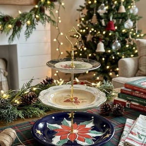 May include: A three-tiered serving stand with gold-colored hardware. The bottom tier is a dark blue plate with a red and green poinsettia design. The middle tier is a cream-colored plate, and the top tier is black with gold accents. Festive Christmas decor.