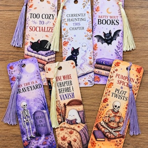 May include: Six rectangular paper bookmarks with rounded corners, each featuring a unique autumn-themed illustration, text, and a decorative tassel. Designs include a black cat, a ghost, a bat, a skeleton, and fall foliage in purple and orange.