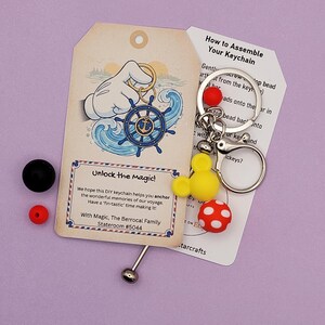 May include: DIY keychain kit with a nautical theme. Includes a tag with a Mickey Mouse hand holding a ship's wheel, a yellow Mickey Mouse head, a red polka dot bead, and a red bead. The tag reads "Unlock the magic!"