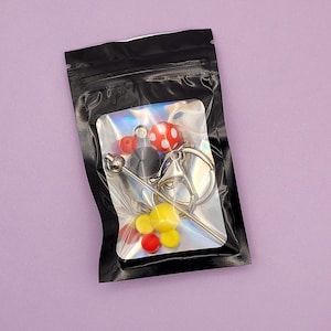 May include: A clear plastic bag containing various craft supplies. The bag holds red and yellow beads, a silver-colored keychain, and a red and white polka-dot bead. The bag is sealed with a black zipper.