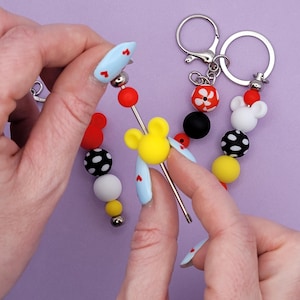 May include: Keychains with colorful beads and charms. One keychain features a yellow Mickey Mouse head charm, red and black beads, and a silver keyring. The other keychain has a white Mickey Mouse head charm and red and black beads.