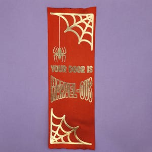 May include: A rectangular red fabric bookmark featuring gold foil accents. The design includes a spider web in the top right and bottom left corners, a hanging spider, and the text "YOUR DOOR IS HARVEL-OUS" printed in a stylized gold font.