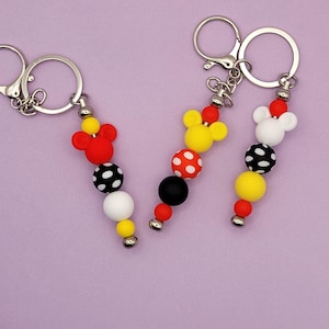 May include: Three keychains with Mickey Mouse-themed designs. Each keychain features a silver ring and a beaded strand with red, yellow, black, and white beads. The top bead is a Mickey Mouse head shape.