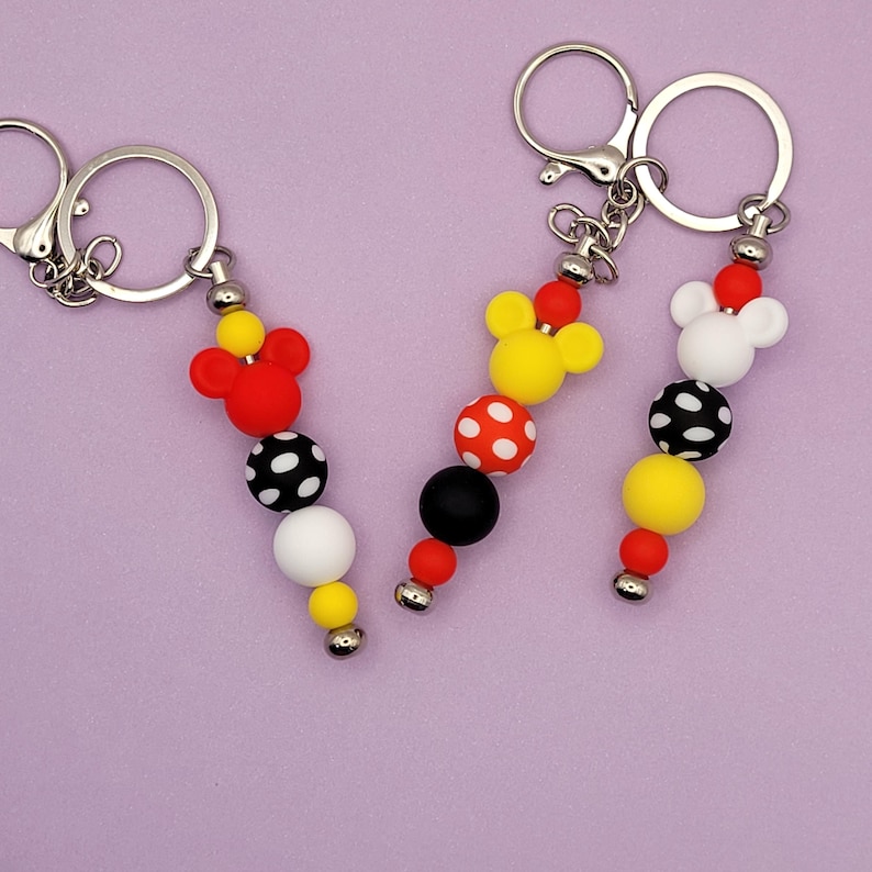 May include: Three keychains with Mickey Mouse-themed designs. Each keychain features a silver ring and a beaded strand with red, yellow, black, and white beads. The top bead is a Mickey Mouse head shape.