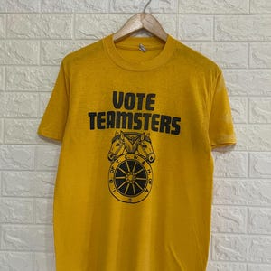 May include: A mustard yellow short-sleeve t-shirt featuring the black text VOTE TEAMSTERS above a graphic of two horse heads and a wagon wheel emblem with the letters B. OF T. C. W. & H. OF A. printed in a circular pattern around the wheel.