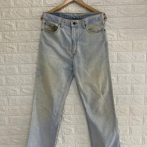 Puede incluir: Vaqueros de mezclilla azul claro con un lavado desgastado, que presentan un diseño clásico de cinco bolsillos, cierre de botones y remaches de color cobre. El pantalón tiene un corte recto y cuelga de una percha de madera sobre una pared de ladrillo.