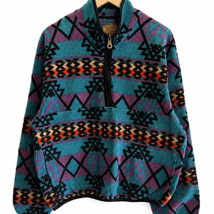 May include: A teal, purple, and black fleece pullover featuring a quarter-zip closure and a geometric pattern with horizontal bands of orange and tan accents. The garment has a high collar, long sleeves, and black trim along the zipper and hem.