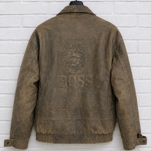 May include: A brown distressed leather jacket featuring a large embossed graphic on the back. The design includes the text SUNTORY COFFEE BOSS and an illustration of a person's face within a circular border. The jacket has a collar and ribbed hem.