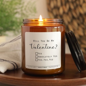 May include: A brown glass candle jar with a white label that reads "WILL YOU BE MY Valentine?" with a checkbox list that says "YES", "ABSOLUTELY YES", and "YES, YES, YES". The candle is lit and the lid is off.