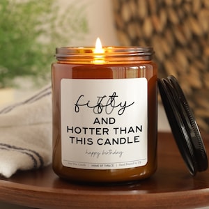 May include: A lit soy wax candle in a brown glass jar with a black lid. The label reads "fifty and hotter than this candle happy birthday". The candle sits on a wooden tray with a white and blue striped cloth.