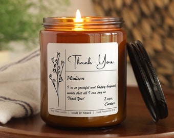 May include: A brown glass candle jar with a white label that says "Thank You Madison I'm so grateful and happy beyond words that all I can say is Thank You! Love, Canter". The candle is lit and the flame is visible.