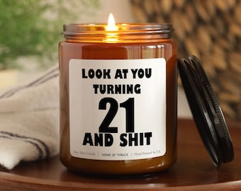 May include: A lit soy wax candle in a brown glass jar with a black lid. The candle has a white label with the text "LOOK AT YOU TURNING 21 AND SHIT". The candle is on a wooden tray.