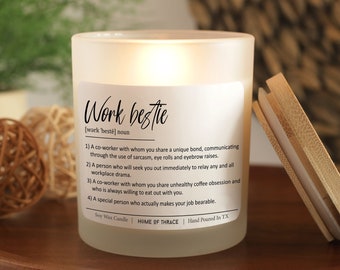May include: A frosted glass candle with a wooden lid, labeled "Work bestie." The label includes a definition of "work bestie" and is hand-poured in TX. The candle sits on a wooden tray with decorative spheres and greenery in the background.