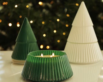 May include: A green glass candle holder with three lit wicks, flanked by two ceramic Christmas tree decorations. One tree is green, the other is white. The candle holder and trees are on a white surface, with a blurred Christmas tree background.