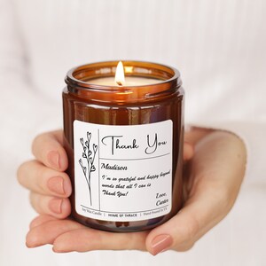 May include: A lit soy wax candle in a brown glass jar. The candle has a white label with the words "Thank You" and a personalized message. The candle is held in a person's hands.