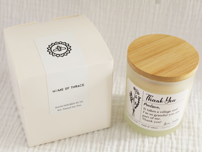 May include: A candle in a glass jar with a wooden lid and a white box. The candle has a label that says "Thank You" and "Madison." The box has a label with a bee design and the words "Home of Thrace."