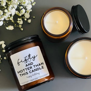 May include: Three soy wax candles in amber glass jars with black lids. One candle has a label that reads "fifty and hotter than this candle happy birthday". The candles are surrounded by white flowers.