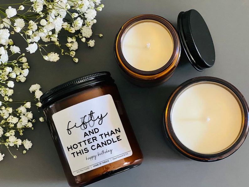 May include: Three soy wax candles in amber glass jars with black lids. One candle has a label that reads "fifty and hotter than this candle happy birthday". The candles are surrounded by white flowers.