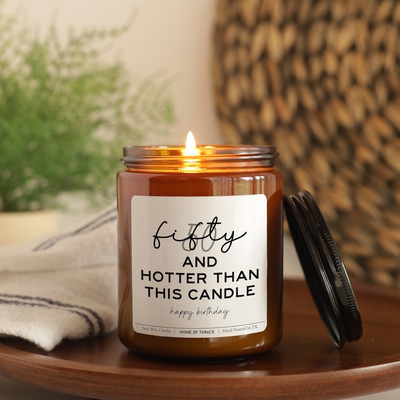 May include: A lit soy wax candle in a brown glass jar with a black lid. The label reads "fifty and hotter than this candle happy birthday". The candle sits on a wooden tray with a white and blue striped cloth.
