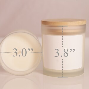 May include: Two frosted glass candles with wooden lids. One candle is viewed from above, showing a 3.0-inch diameter. The other candle is 3.8 inches tall and has a blank white label. The candles are on a light pink surface.