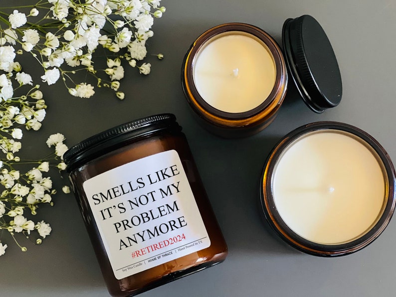 May include: Three amber glass candle jars with white wax and black lids. The centre jar has a white label with the text "Smells like it's not my problem anymore #RETIRED2024".