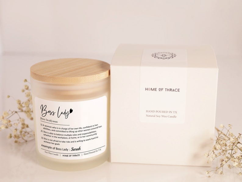 K&ouml;nnte beinhalten: Ein mattes Glas-Kerzengef&auml;&szlig; mit einem Holzdeckel. Die Kerze ist mit "Boss Lady" beschriftet und enth&auml;lt eine Definition des Begriffs. Die Kerze befindet sich in einer wei&szlig;en Box mit dem Text "Home of Thrace" und "Hand Poured in TX".