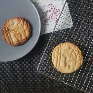 Op de afbeelding: Twee suikerkoekjes met een boslandschapdesign. De koekjes liggen op een grijs bord en een draadrek. De koekjes zijn lichtbruin van kleur en hebben een licht gouden tint. Het ontwerp op de koekjes is een silhouet van een hert in een bos met bomen en een maan.