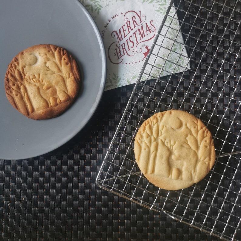 Op de afbeelding: Twee suikerkoekjes met een boslandschapdesign. De koekjes liggen op een grijs bord en een draadrek. De koekjes zijn lichtbruin van kleur en hebben een licht gouden tint. Het ontwerp op de koekjes is een silhouet van een hert in een bos met bomen en een maan.