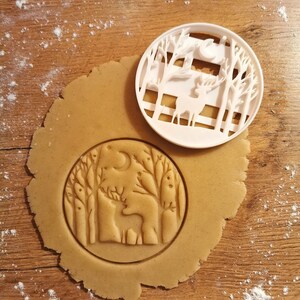 Op de afbeelding: Een witte plastic koekjesvormer in de vorm van een hert dat in een bos staat met bomen en een halve maan. De koekjesvormer ligt op een rond stuk koekdeeg met hetzelfde ingegraveerde ontwerp.
